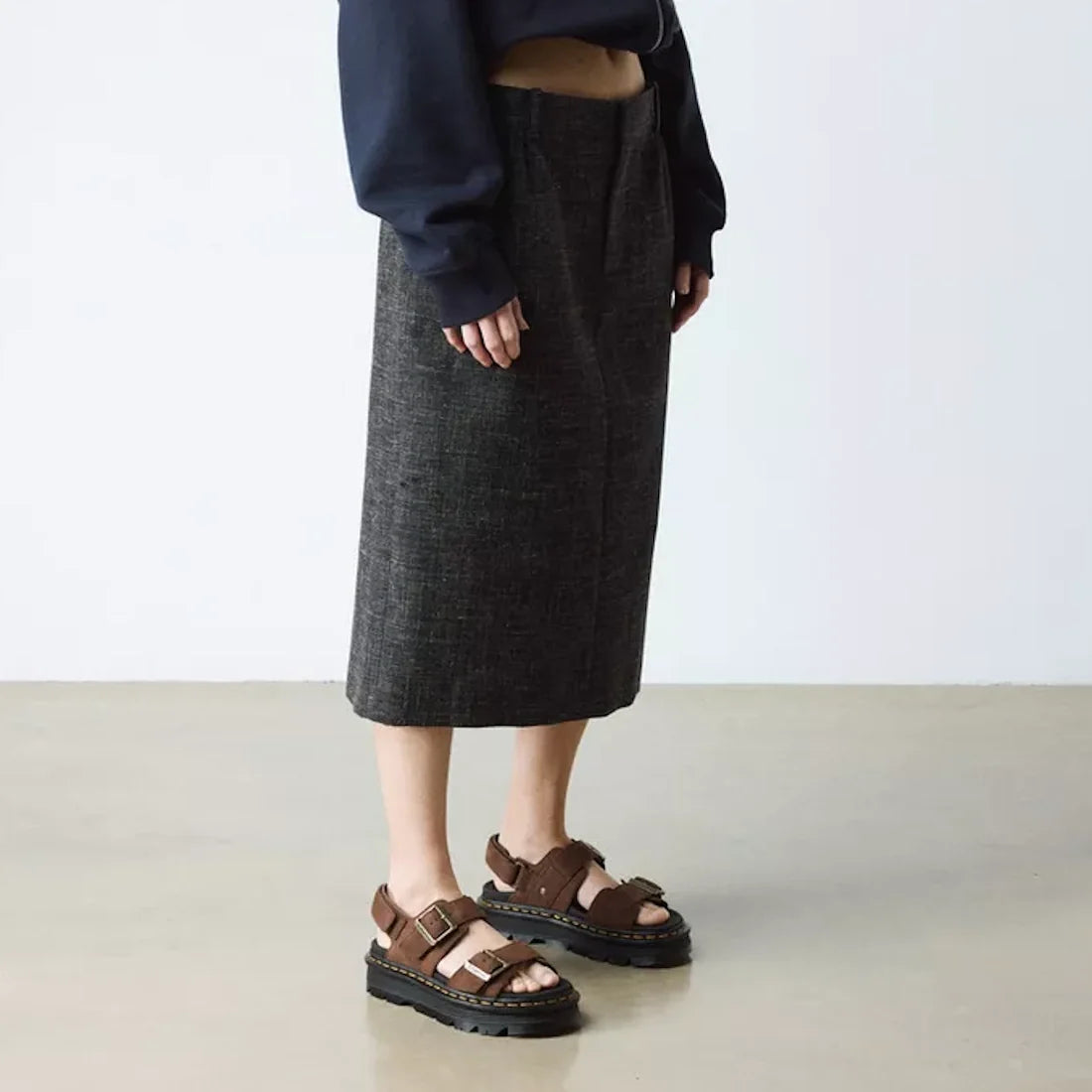 Indoors, a person wears a dark gray knee-length skirt, navy long-sleeve top with extended sleeves, and Dr Martens Zebzag Sepia Brown Nubuck 2 Strap Sandals. The minimal background features a light floor and wall., 190665790603, 190665790610, 190665790627,
