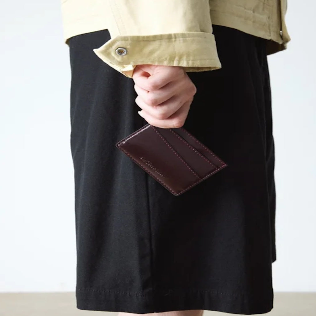 A person wearing a light-colored jacket and black skirt holds the Dr Martens Cardholder II Cherry Red Oxford Brush Vegan in their right hand, with their arm resting by their side.