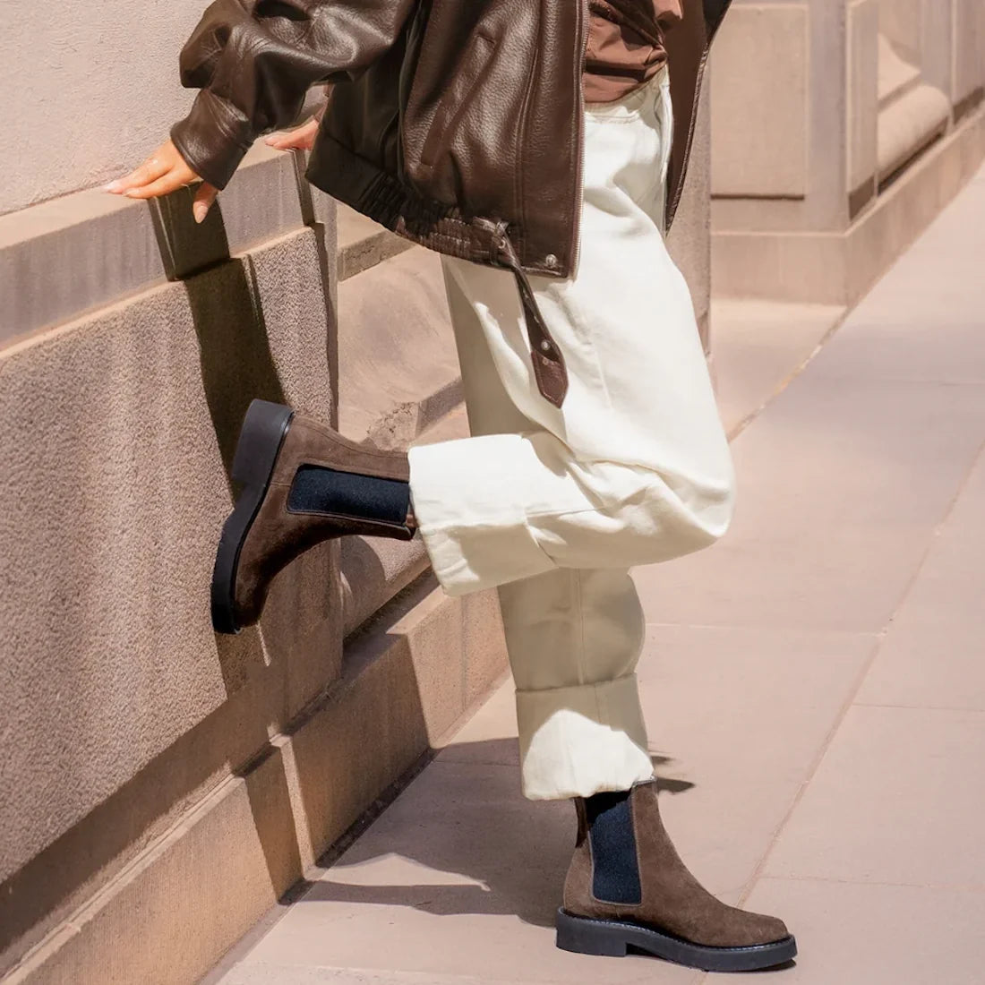 A person in light pants, a brown leather jacket, and EOS Womens Leilin Suede Kalamata Brown Ankle Pull On Boots stands on the sidewalk, one foot raised and resting against a stone building wall., 9321190037555, 9321190037562, 9321190037579, 9321190037586,