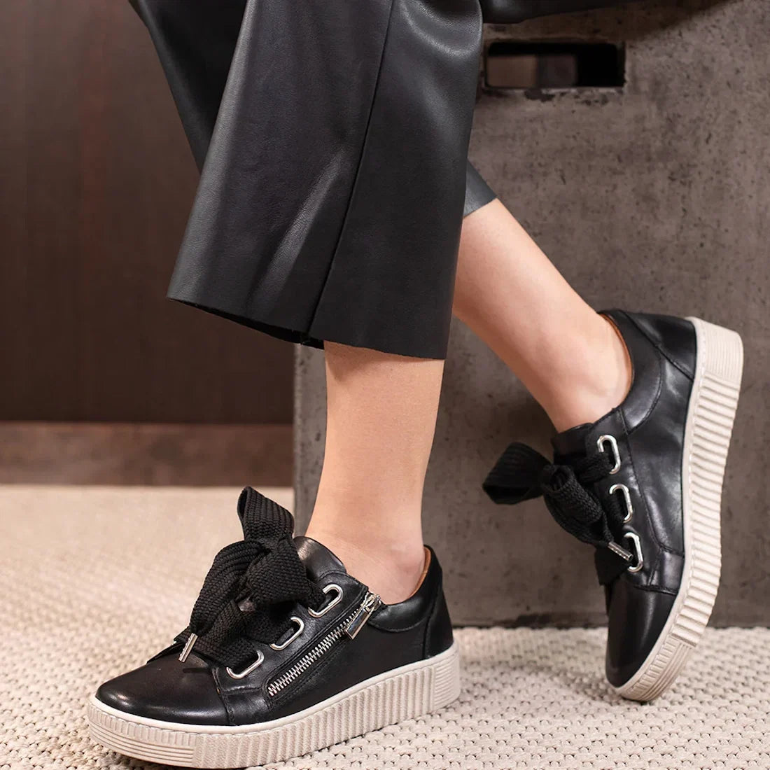 A person wearing black leather pants and EOS Joyce Leather Trainer Black Lace Up with Zip sneakers, featuring thick soles, large black laces, anatomical insoles, and side zippers, rests their feet on an indoor concrete surface.