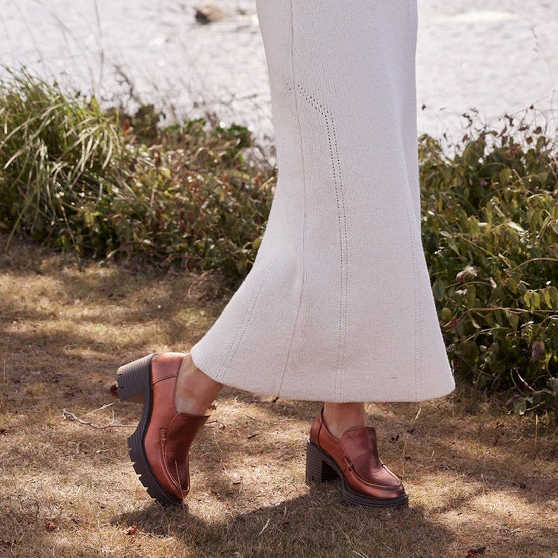 A person in a long, light skirt stands outdoors on dry grass near greenery, showcasing EOS Malia Rusty Metallic Heeled Leather Shoes., 36555, 9321191196909, 9321191196916, 9321191196923, 9321191196930, 9321191196947, 36561