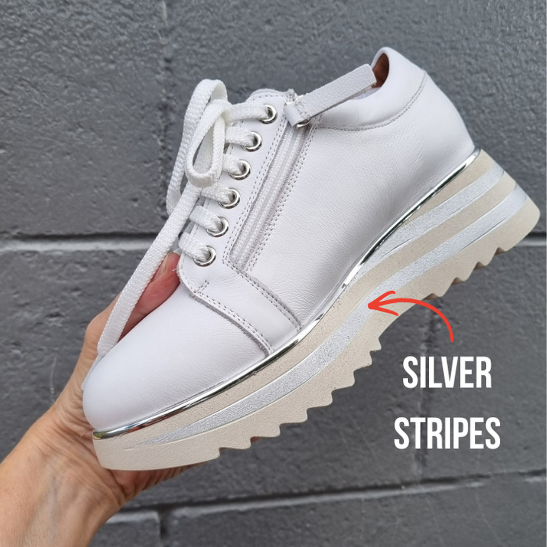 A hand holds the Alfie & Evie Hosting Womens Leather Shoe Platform White/Silver, featuring white laces, a zipper, and silver metallic stripes on the midsole. "SILVER STRIPES" text with an arrow highlights the detail against a gray brick wall., 43583, 4358