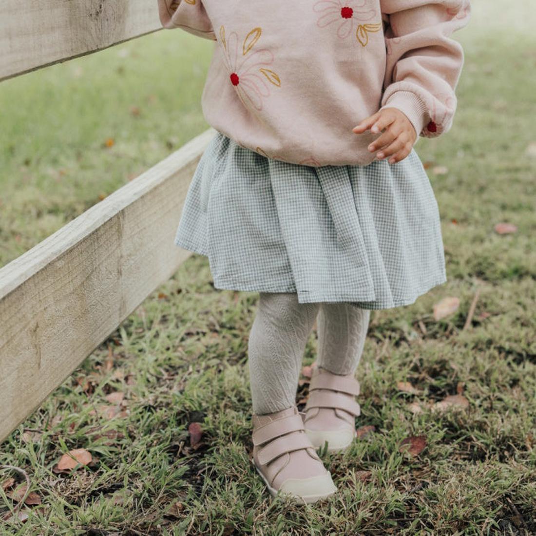 A young child’s lower body is visible, wearing a pink flower sweatshirt, blue checkered skirt, white tights, and Pretty Braver Blake Leather Boot First Walker Blush shoes on grass near a wooden fence., 9400275329195, 9420075329188, 9420075329171