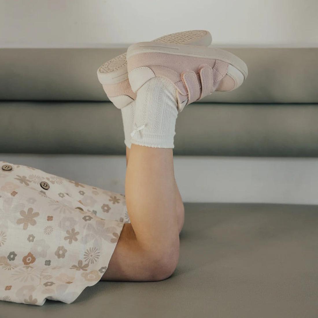 A child in a floral dress, white socks, and Pretty Brave Otto Organic Canvas Trainer With Leather Toe Buffer Pink lies on their back with legs raised, resting on a gray cushioned surface., 9420075314719, 9420075314702, 9420075316157, 9420075314696, 942007