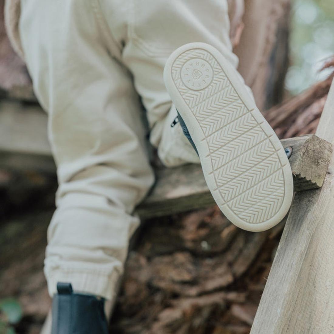 A person in light-colored pants and Pretty Brave Electric Leather Boot - Jet Black sits outdoors on a wooden structure, showing the shoe’s lightning bolt sole design to the camera., 35922, 9420075327986, 35924, 35925, 9420075327955, 9420075327948, 9420075