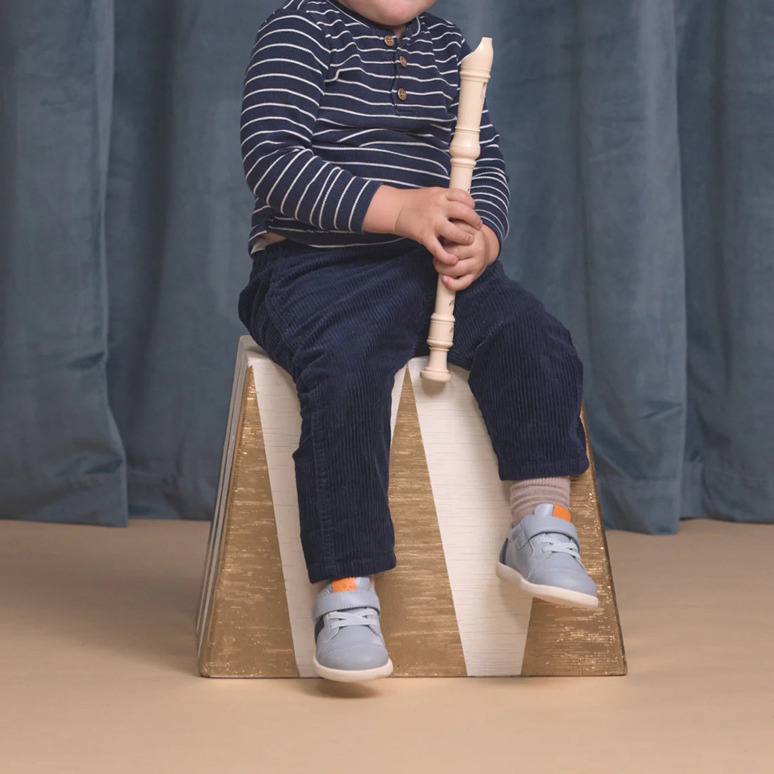 A young child, wearing a navy striped shirt, blue pants, and Pretty Brave SID Leather Trainer First Walker Indigo Blue Grey shoes, sits on a gold and white block holding a white recorder; the child's face is not visible., 9420075362918, 9420075362901, 942