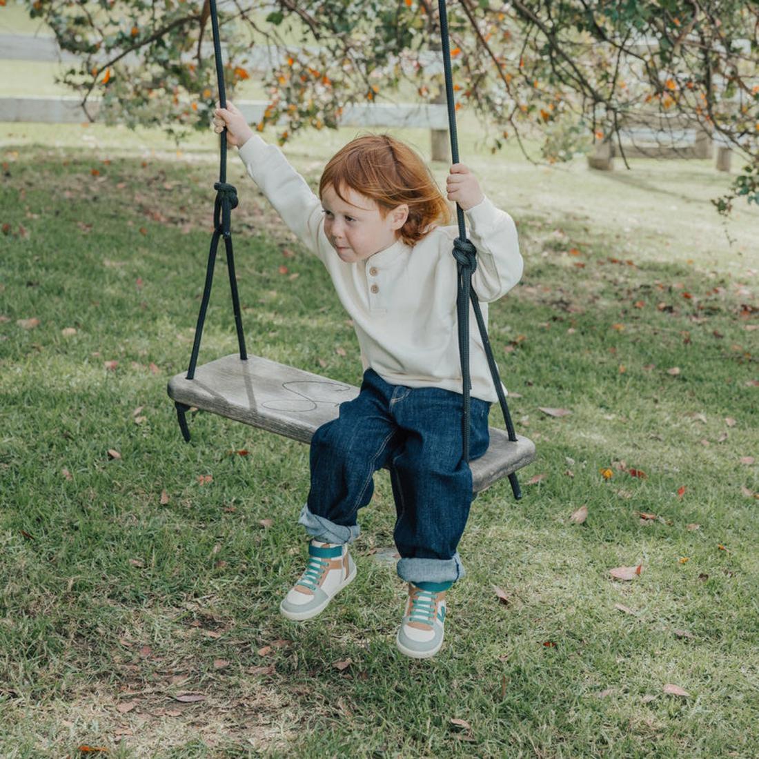 A young child with red hair, in a white sweater and jeans, wears Pretty Brave Hi Top Boot Forest Mix for a supportive fit while sitting on a swing outdoors amid trees and fallen leaves., 35902, 35903, 35904, 35905, 35906, 35907, 35908, 9420075328709, 3591