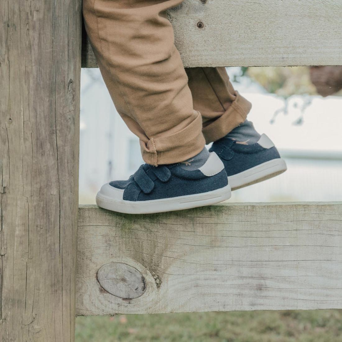 A child, with only legs and feet visible, wears tan pants and Pretty Brave Otto Organic Canvas Trainer With Leather Toe Buffer Black while climbing or sitting on a wooden fence., 35870, 35871, 9420075329584, 9420075329577, 9420075329560, 5420055329553, 94