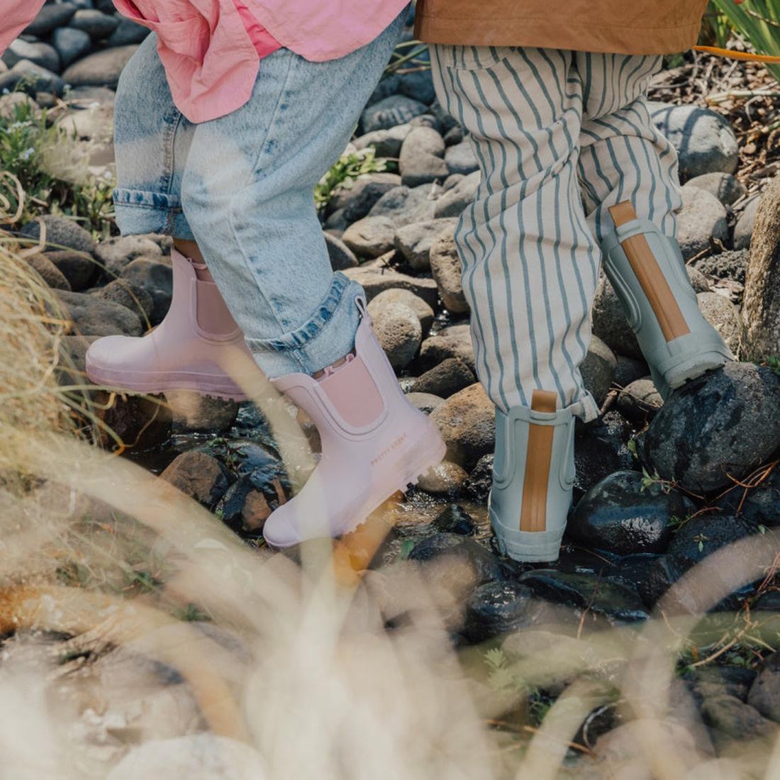 Two children wear casual clothes and stand on wet rocks outdoors. One wears Pretty Brave Puddle Boot Ankle Rainboot Blush Pink and jeans, while the other has gray boots with striped pants. Green plants grow among the rocks., 9420075328525, 9420075328518,