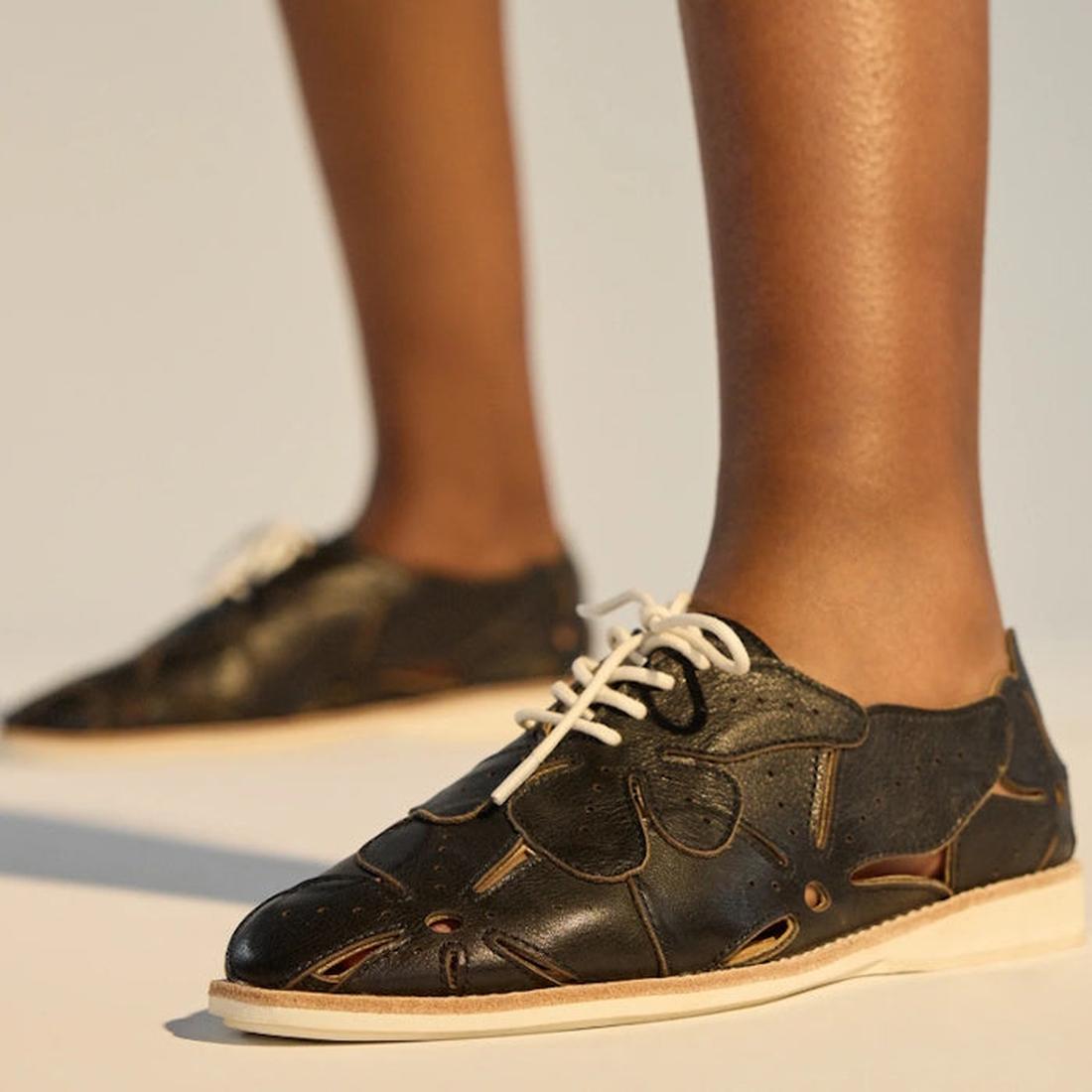 Close-up of a person wearing Rollie Derby Applique Black Womens Leather Brogue Shoes with cut-out soft leather uppers and beige soles, standing on a light surface. Only the lower legs and feet are visible., 9781760792312, 9781760792329, 9781760792336, 978