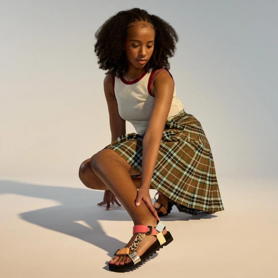 A woman with curly hair kneels on one knee, wearing a sleeveless white top, green plaid skirt, and the Rollie Womens Sandal Tooth Wedge Leopard Multi with colorful adjustable leather straps against a neutral background., 9339554333786, 9339554333793, 9339