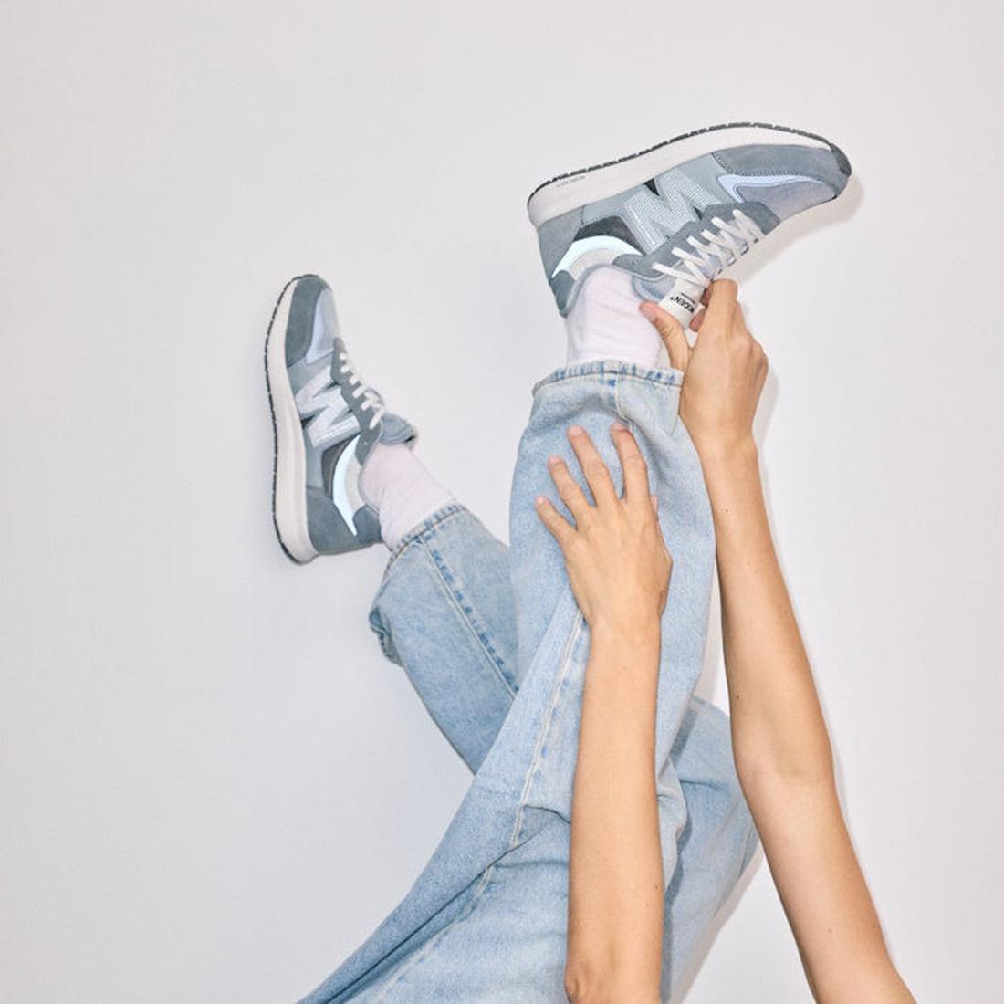 A person in light blue jeans and Woden Rigmor Reflective Women's Sneaker Grey Multi with white laces lifts one leg with both hands against a plain, light-colored background., 43563, 5713326601046, 5713326601053, 5713326601060, 43567