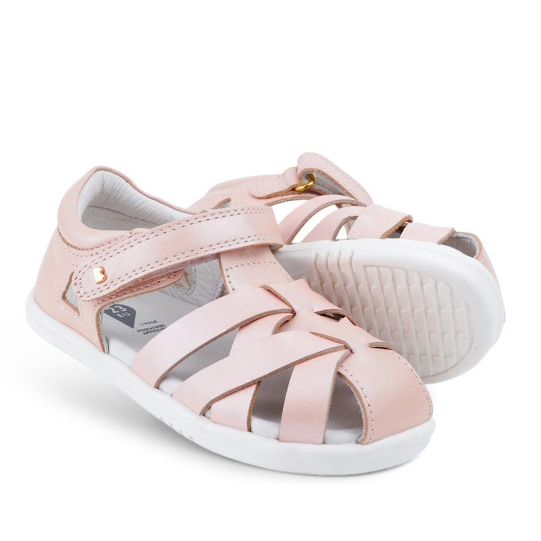 The Bobux Iwalk IW Tropicana II Sandal 638305 in Seashell Pink features crisscross straps, white soles, and a Velcro closure for toddlers. One sandal is upright while the other leans against it, showcasing this stylish summer shoe.