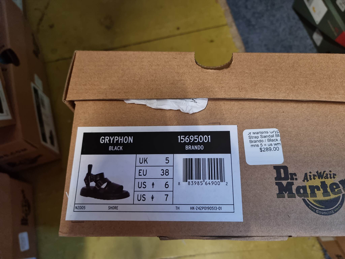 A close-up of a Dr Martens Gryphon Strap Sandal Black Brando box label shows sizes UK 5, EU 38, US 6 (women), US 7 (men), and a $289.00 price. Part of the box and floor are visible beneath this gladiator-inspired sandal's details., 883985648982, 883985648