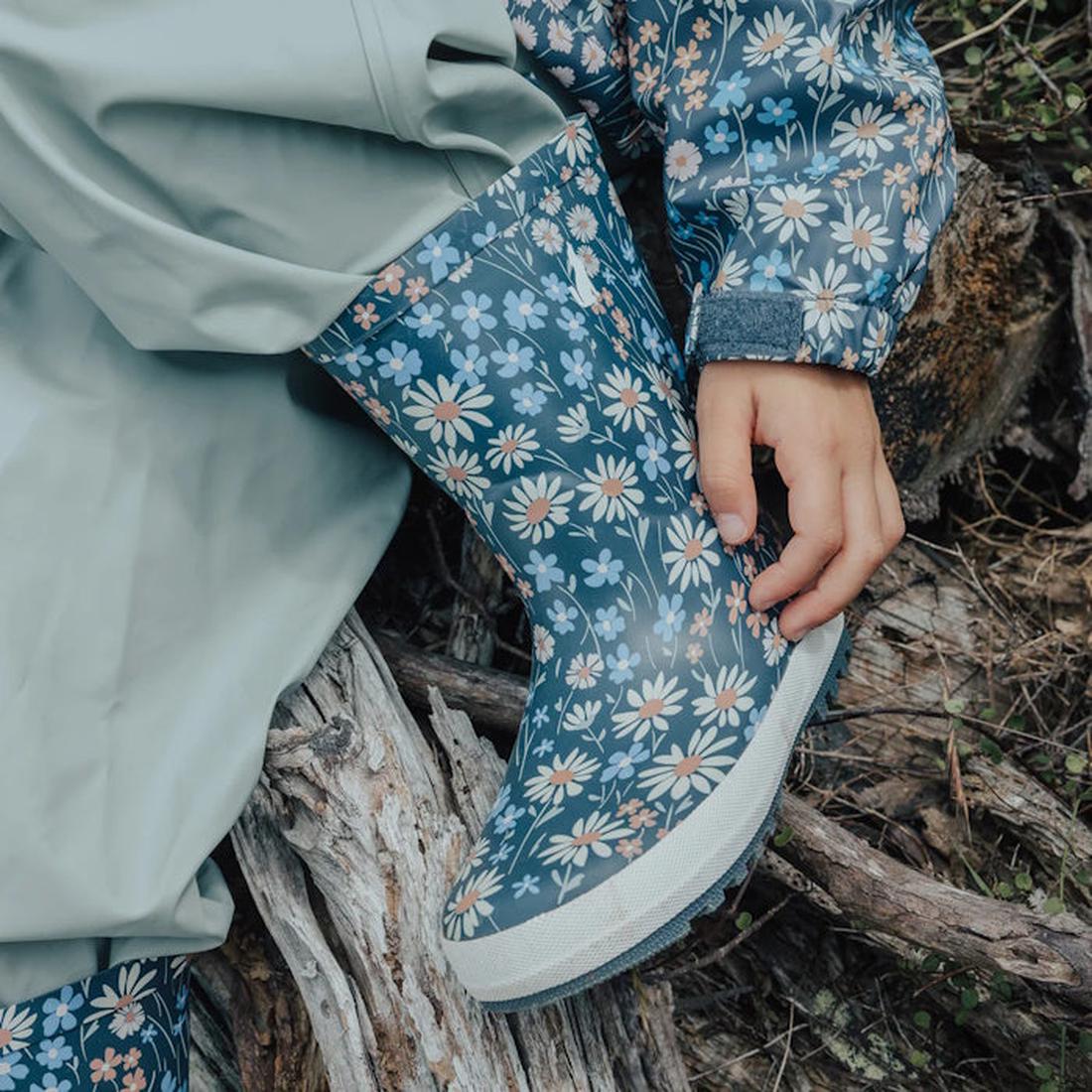 A child wearing a floral-patterned raincoat and the CryWolf Rain Boots Winter Floral Navy sits on a log, touching their boot with one hand.