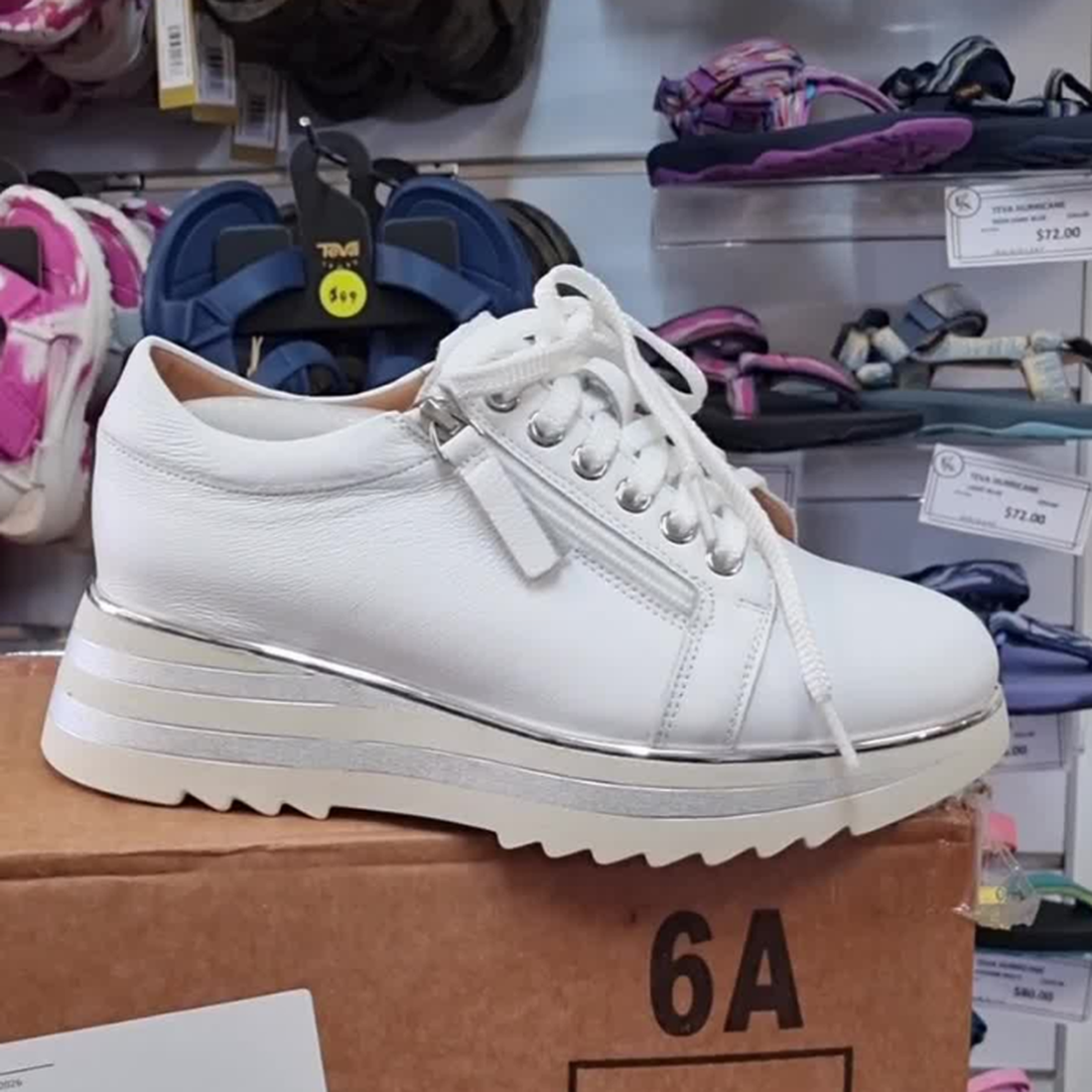 The Alfie & Evie Hosting Womens Leather Shoe Platform White/Silver is displayed on a box in a shoe store among sandals and price tags. This stylish sneaker features silver details, a side zipper, and offers lightweight comfort., 43583, 43584, 43585, 43586