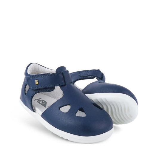 The Bobux Step Up Zap II Navy Closed Toe Sandal 732404 Quick Dry features navy blue uppers, white soles, a closed-toe with cut-out shapes, and an adjustable ankle strap. Both side and sole are visible in the angled display.