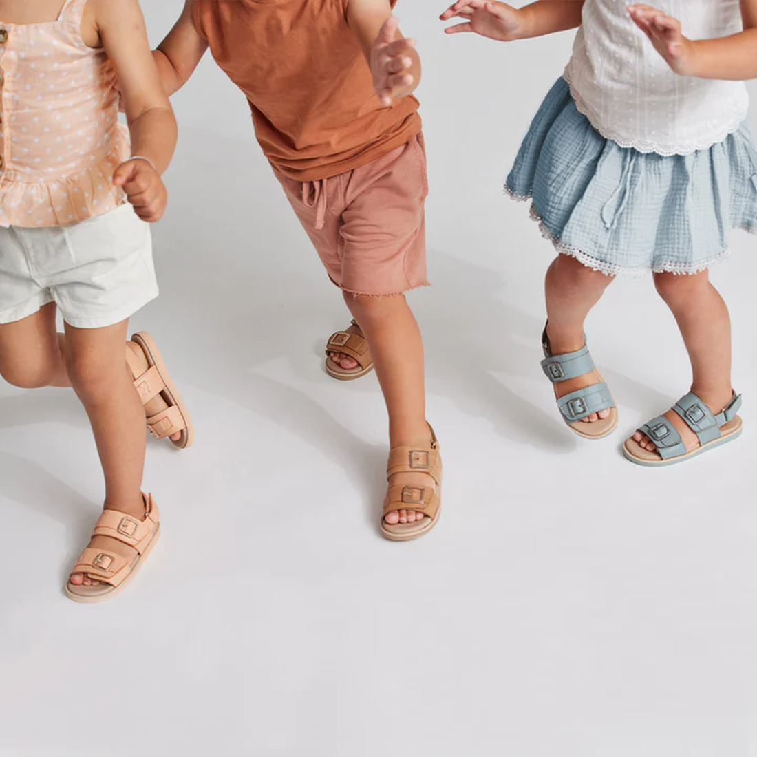 Three children in summer outfits walk against a white background. Their faces are unseen, and they wear Pretty Brave Alex Leather Sandal Coral (sizes 29-33), featuring adjustable velcro straps for a secure fit. CLEARANCE NOT RETURNABLE., 9421036357554, 94