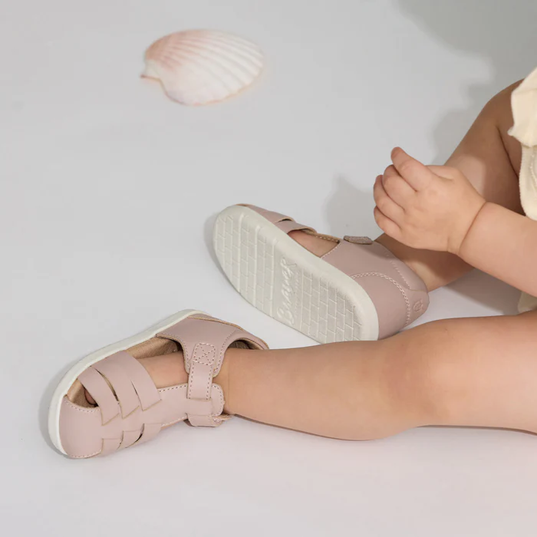 A small child sits on a white surface wearing the Pretty Braver Billie First Walker Sandal Closed Back in Blush, with a seashell nearby. The child's legs are visible as they adjust or play with their hands., 9420075334458, 9420075334441, 9421036357110
