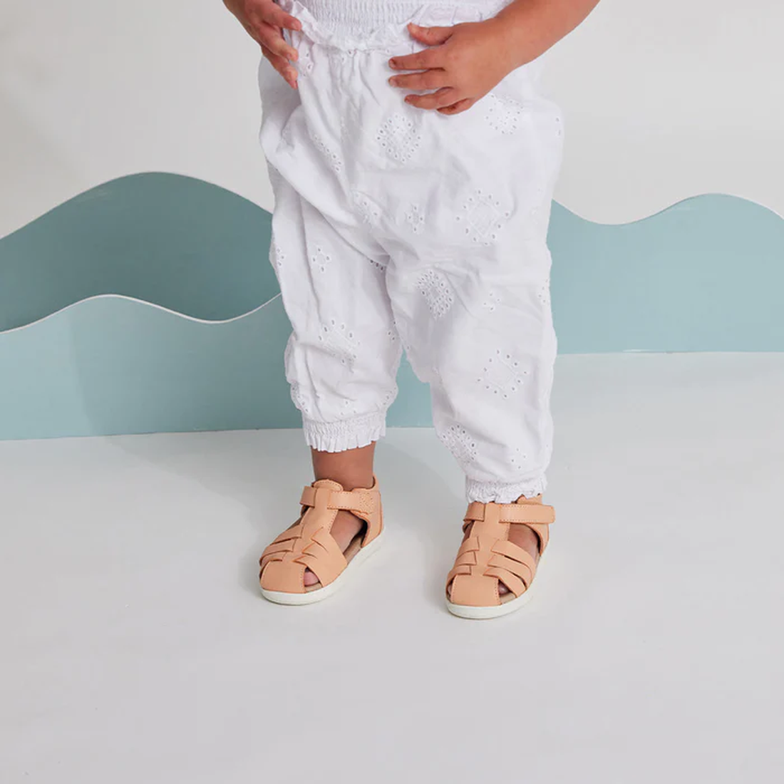 A toddler wearing white eyelet pants and Pretty Braver Billie First Walker Sandal Closed Back Coral, a lightweight flexible sandal for early walkers, stands on a white floor with a light blue wavy backdrop. The child's hands are near their waist., 9421036