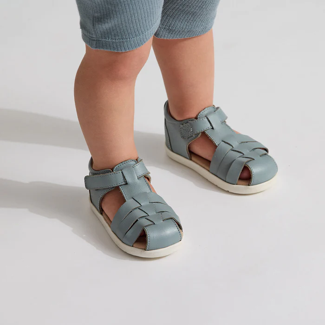 A toddler wears Pretty Braver Billie First Walker Sandal Closed Back Seafoam, ideal for early walkers, with light blue ribbed pants while standing on a white surface., 9421036357202, 9421036357219, 9421036357196