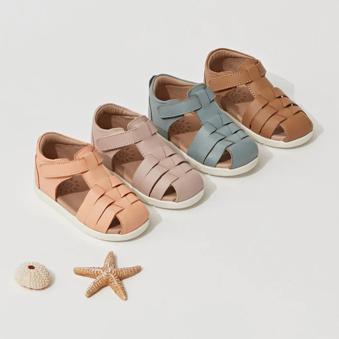 Four pairs of toddler sandals, including the Pretty Braver Billie First Walker Sandal Closed Back in Seafoam, are arranged on a white surface with seashell and starfish accents—ideal for your early walker’s summer adventures., 9421036357202, 9421036357219