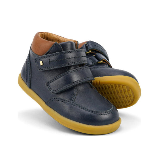 The Bobux Iwalk Timber Navy Leather Boot 632619 features premium dark blue leather, tan soles, brown collars, and double adjustable straps—one boot stands upright as the other leans against it.