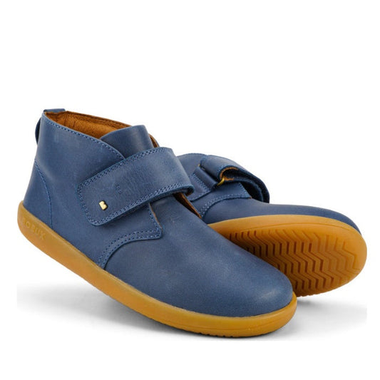 The Bobux Kid+ Desert Boot Midnight Blue 830312 is a pair of unisex blue leather high tops with tan rubber soles, featuring a wide velcro opening and minimalist design. One boot stands upright while the other lays on its side.