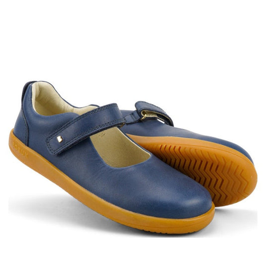 The Bobux Kid+ Delight Mary Jane Navy 831615Z features classic navy blue leather, a Velcro strap, and light brown rubber soles. One shoe stands upright, the other lies on its side, displaying the tread pattern.