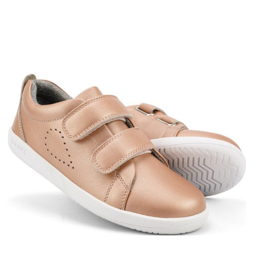 The Bobux Kids + Plus Grass Court Rose Gold Velcro Trainers 832455 feature a leather upper, white soles, double velcro straps, and small side perforations. One shoe stands upright while the other leans against it.