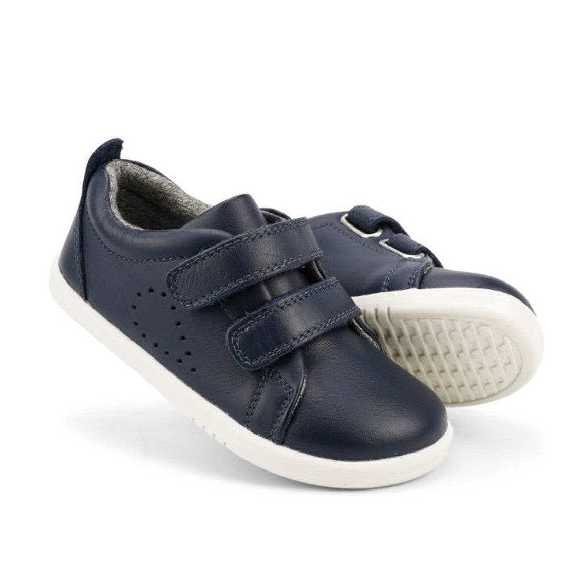 The Bobux Iwalk Grass Court Navy Leather Trainer 633723J kids' shoes feature navy leather, white soles, and two Velcro straps. Displayed on a white background, one shoe stands upright while the other lays on its side to show the textured sole.