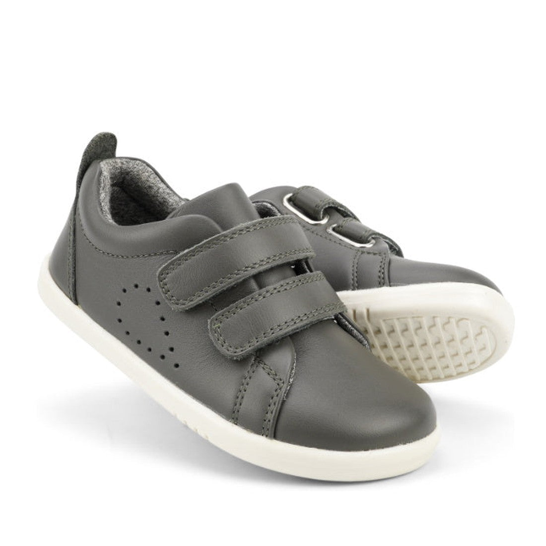 The Bobux Iwalk Grass Court Smoke Grey Leather Trainer 633702 is a unisex kids' sneaker with white soles, dual Velcro straps, and side perforations for ventilation—shown upright and tilted to reveal the sole.