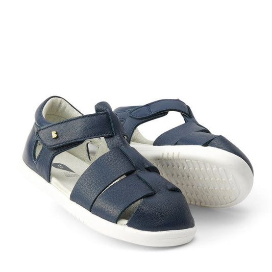 The Bobux IWalk Tidal Closed Toe Quick Dry Sandal Navy 634404A features a navy blue design, white rubber soles, velcro strap, cutout details, and quick dry leather, shown against a white background.