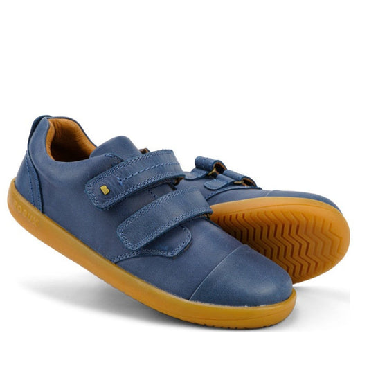 The Bobux Kid+ Port Shoe Midnight Blue 833007 features a blue leather upper, tan rubber soles, and two Velcro straps. One shoe stands upright while the other is tilted to display the textured sole.