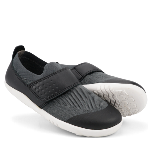 A pair of black Bobux Kid+ Lo Dimension III slip-on shoes (838604 CLEARANCE), featuring Velcro straps and flexible soles; one shoe stands upright while the other lies on its side to display the lightweight sole.