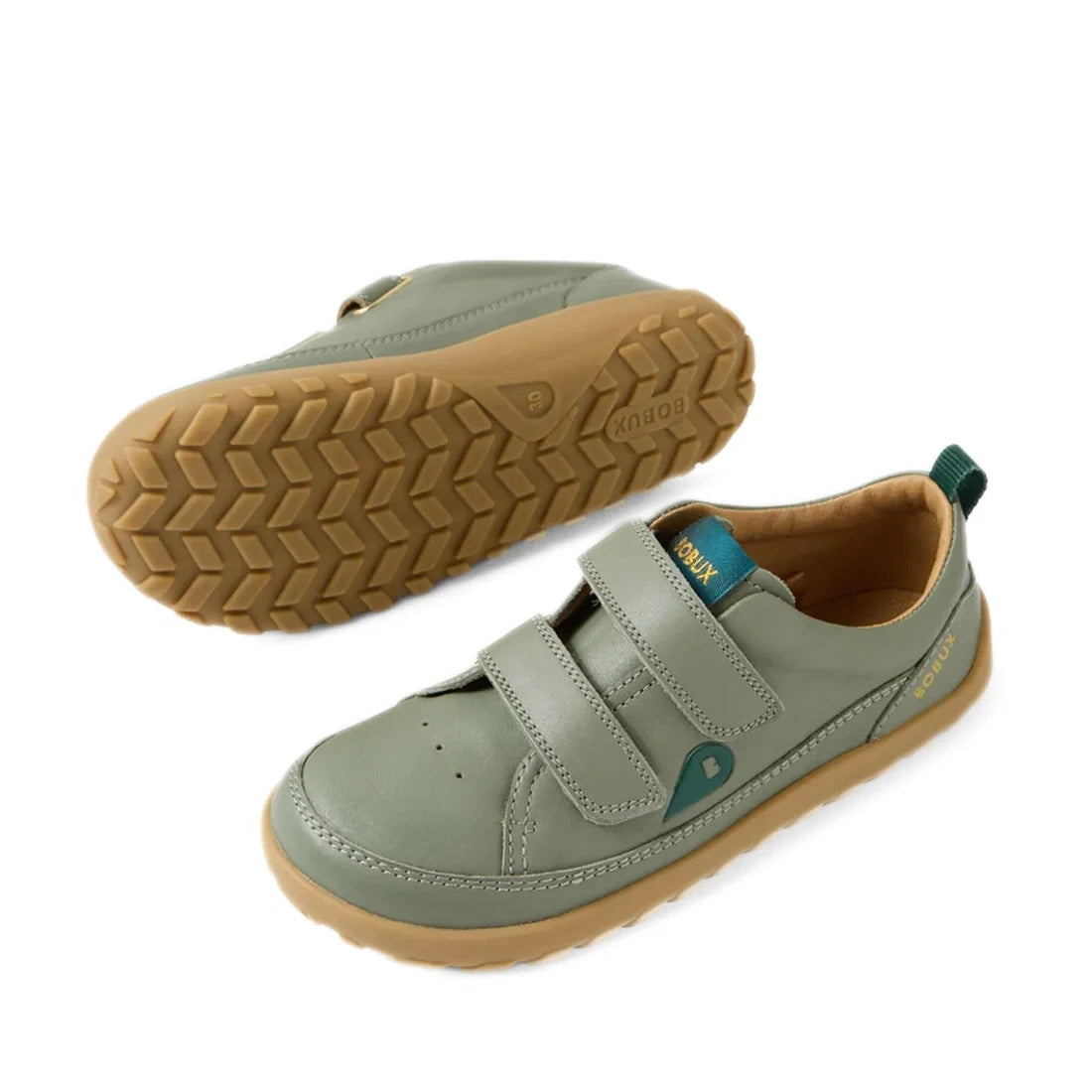 The Bobux KP Kids Plus Dawn Leather Sneaker in Vetiver Green features a barefoot-inspired design with tan rubber zero-drop soles, two Velcro straps, and a teal pull tab at the heel. One sneaker is shown upright while the other displays its tread pattern a