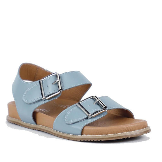 The Bay Lane Zeus Denim Blue Women's Leather Sandal with Back Strap features a light blue color, two adjustable silver buckles, open toe, closed heel, visible stitching on the flat brown sole, and a Luxe Support footbed for comfort.