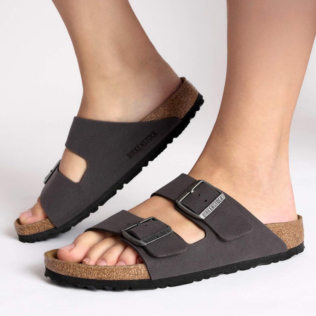 Someone stands on a white background wearing Birkenstock Arizona Velvet Grey Birkibuc – Regular Fit sandals, featuring two adjustable buckle straps and a supportive cork-latex footbed., 48705, 48706, 4066648916567, 4066648916604, 4066648916642, 4066648916