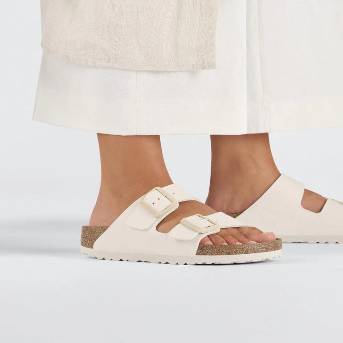 Beige Eggshell Birkenstock Arizona Double Strap  sandals worn with white pants on a light gray background.,
4066648369509,
4066648369547,
4066648369585,, 46716, 46717, 4066648369509, 4066648369547, 4066648369585, 46721, 46722, 46723, 46724, 46725, 46726, 