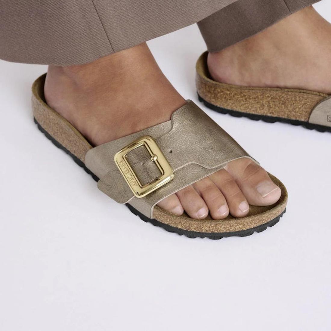 Close-up of a person in brown pants wearing Birkenstock Womens Catalina Cushion Buckle Birko Flor Graceful Taupe sandals (regular width) with a large gold buckle, cork-latex footbed, and black outsoles, standing on a light surface., 4067707018482, 4067707