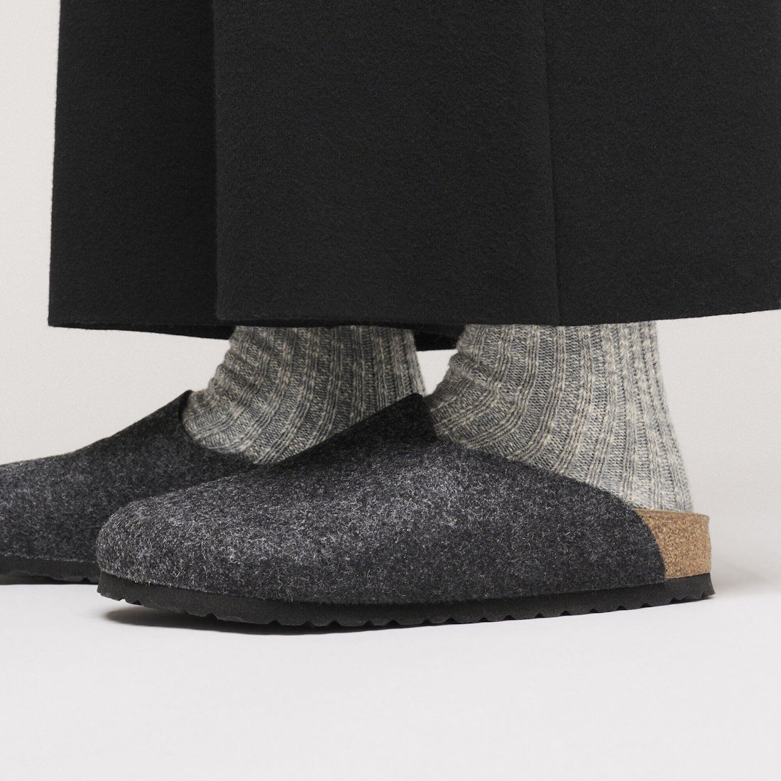 Close-up of a person wearing Birkenstock Amsterdam Felt Wool Clog in Anthracite Grey (Regular Width) with thick light gray ribbed socks and black pants, showcasing the supportive cork footbed and stylish, comfortable design., 52004, 52005, 4067707295241, , 4067707295289, 4067707295326, 4067707295364, 4067707295401, 4067707295449, 4067707295487