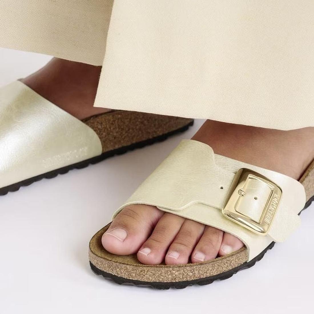 Close-up of a person wearing Birkenstock Womens Catalina Cushion Buckle Birko Flor Pearl White sandals (Regular Width) with a signature cork-latex footbed and large gold buckle, paired with light pants on a white surface., 4066648995265, 4066648995302, 40