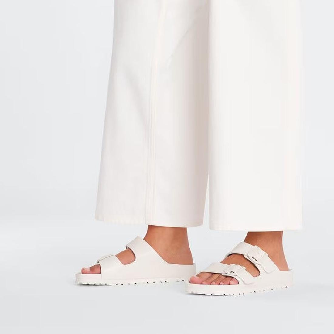 A person wears wide-leg white pants and Birkenstock Arizona EVA Eggshell Off White Narrow Width sandals, standing against a plain light background with only their lower legs and feet visible., 47533, 47534, 4066648352167, 4066648352204, 4066648352242, 406
