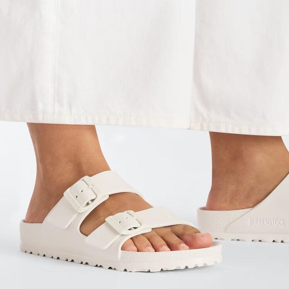 A person stands on a white floor wearing Birkenstock Arizona EVA Eggshell Off White Narrow Width sandals with two buckle straps. Only their lower legs and feet in white pants are visible., 47533, 47534, 4066648352167, 4066648352204, 4066648352242, 4066648