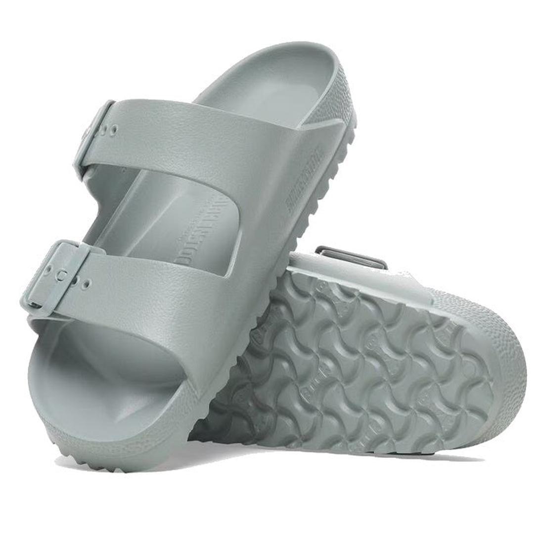 A pair of Birkenstock Arizona EVA Pure Sage Green Narrow Width sandals with two adjustable straps and textured soles. One stands upright while the other lies on its side, showing the signature sole pattern inspired by the Arizona design.