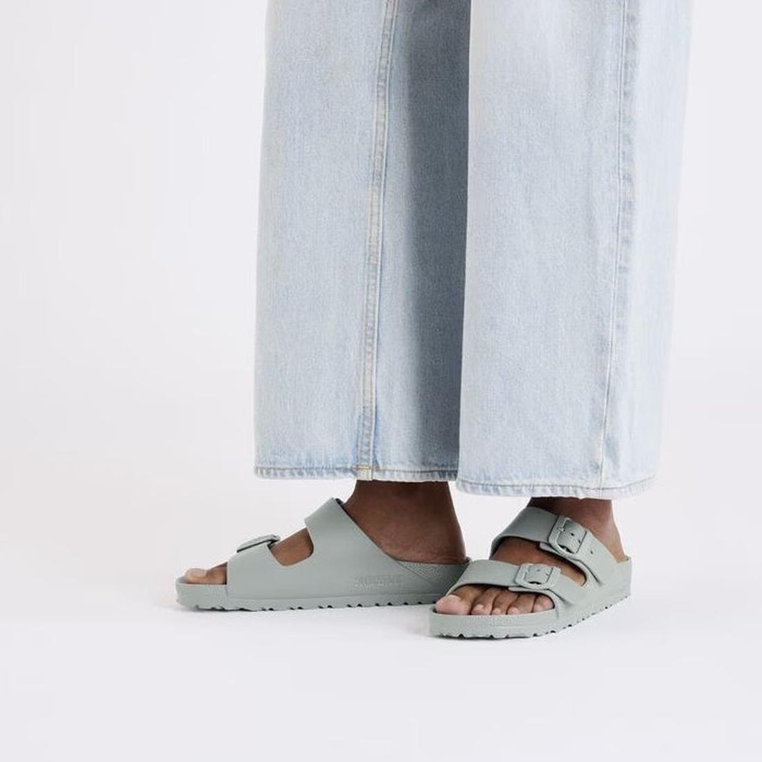 A person wearing light blue wide-leg jeans and Birkenstock Arizona EVA Pure Sage Green Narrow Width sandals stands against a plain white background., 47527, 4067707110742, 4067707110780, 4067707110827, 4067707110865, 4067707110902