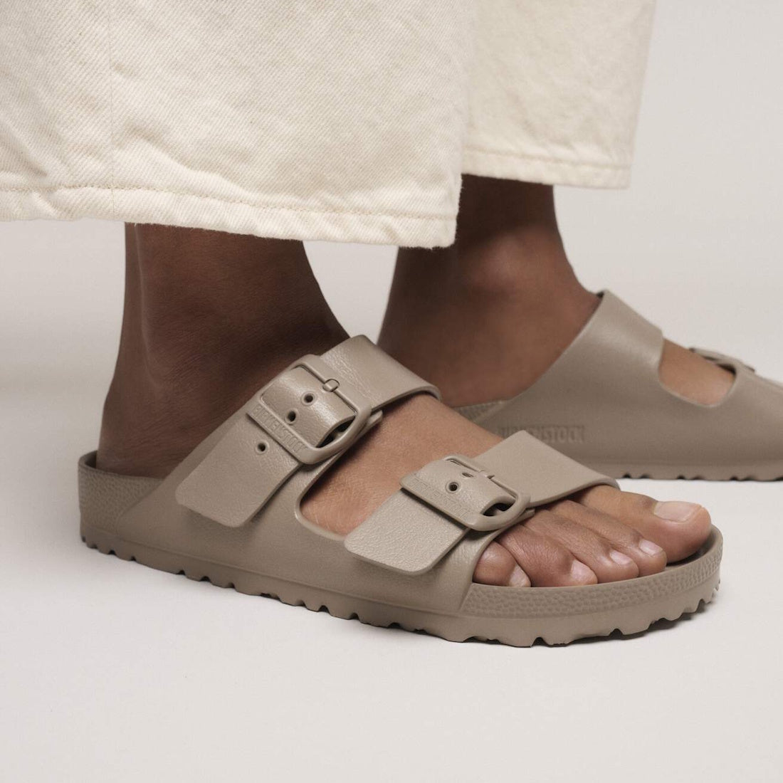 Close-up of a person wearing Birkenstock Arizona EVA Grey Taupe Regular Fit sandals with large buckles, styled with off-white wide-leg pants. The waterproof slides stand out against a plain, neutral background., 4067707373499, 4067707374939, 4067707374977, 4067707375011, 4067707375059