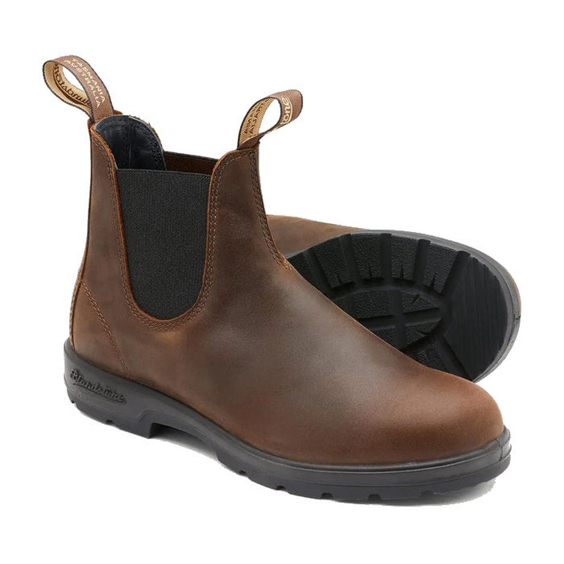 The Blundstone 1609 Chelsea Boots in antique brown leather feature black elastic side panels, pull tabs, and water-resistant material. Their sturdy black soles with visible tread patterns enhance durability. One boot stands upright, while the other lies o