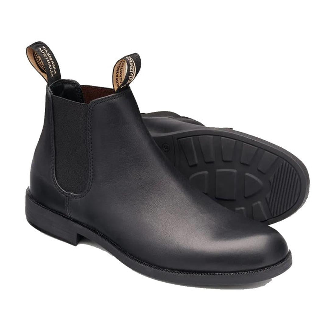 A pair of Blundstone 1901 Mens Dress Black Boots, tapered fit with pull tabs and elastic side panels, is shown on a white background. One boot stands upright; the other reveals its treaded sole. Versatile ankle boots for any occasion.