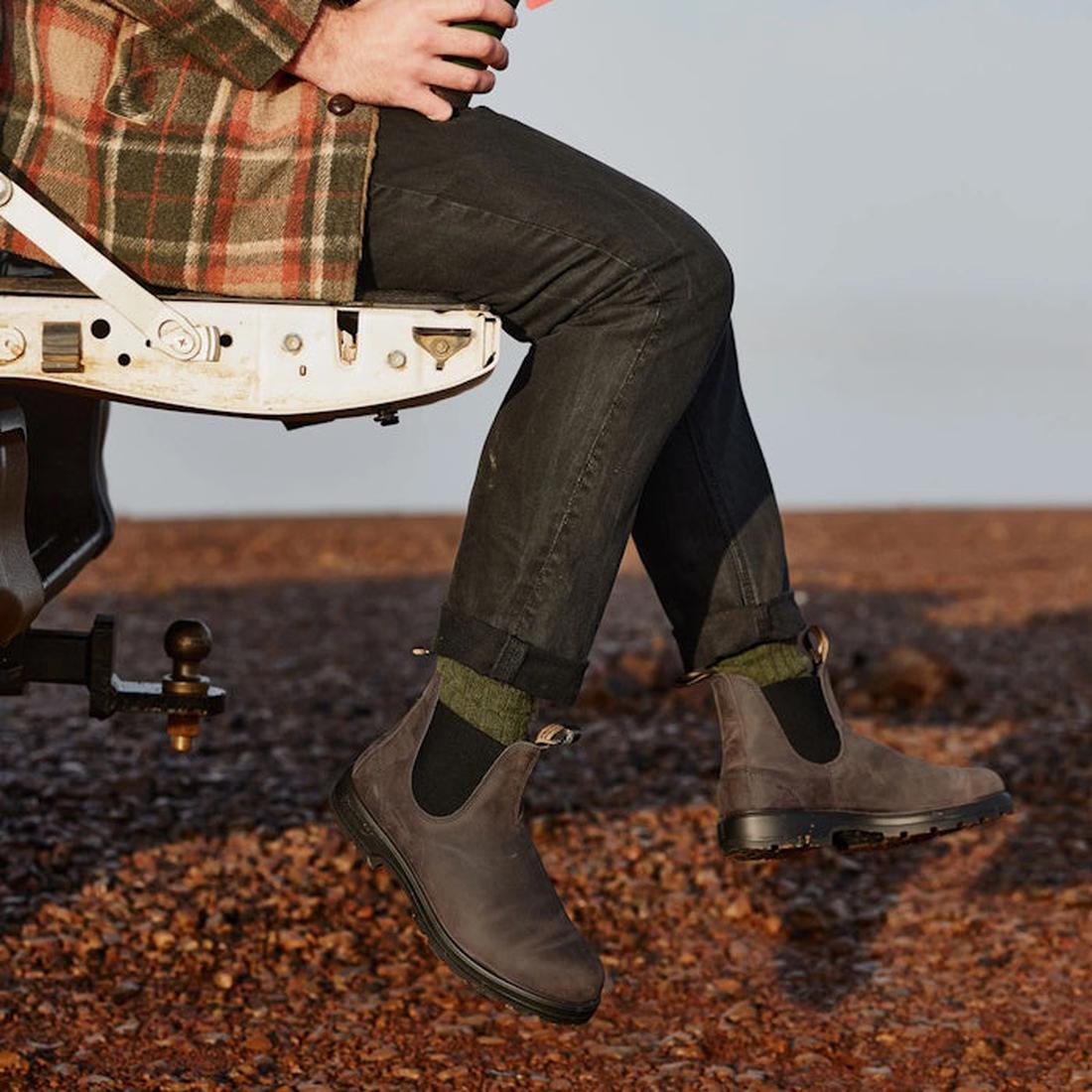 A person in a plaid jacket, dark pants, green socks, and Blundstone 2446 Chelsea Boots in Vintage Clay Brown sits on an open vehicle tailgate, holding a mug with vintage charm against rocky ground., 9315891575034, 9315891575041, 9315891575058, 93158915750